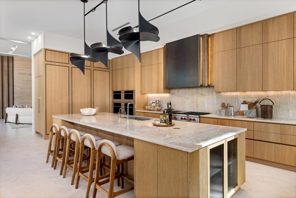 Spectrum’s stevali luxury kitchen featuring light rift-cut oak cabinetry with integrated appliances, a large marble island with ribbed detailing, and fluted glass display cabinets.. Three full-height doors with metal inlay hide a pantry. A custom black and gold ombre hood is visible over the range, and unique black sculptural pendant lights hang above the island.