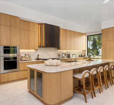 Italian kitchen with rift-cut oak cabinetry, a large island with ribbed detailing, fluted glass doors for display cabinets, integrated seating, stainless steel double ovens, and a custom hood in an ombre finish. A large window offers a view of lush greenery.