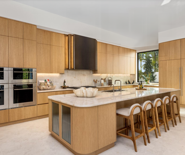 Panther National Spec Home: Italian Kitchen by Spectrum Fine Cabinetry Italian kitchen with rift-cut oak cabinetry, a large island with ribbed detailing, fluted glass doors for display cabinets, integrated seating, stainless steel double ovens, and a custom hood in an ombre finish. A large window offers a view of lush greenery.