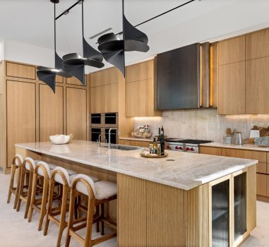 Spectrum’s stevali luxury kitchen featuring light rift-cut oak cabinetry with integrated appliances, a large marble island with ribbed detailing, and fluted glass display cabinets.. Three full-height doors with metal inlay hide a pantry. A custom black and gold ombre hood is visible over the range, and unique black sculptural pendant lights hang above the island.